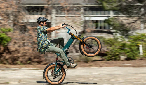 Un cycliste réalise une roue arrière sur un vélo électrique à pneus larges Fiido M1 Pro, mettant en valeur son cadre pliant compact, ses pneus larges et sa suspension avant sur une route urbaine.