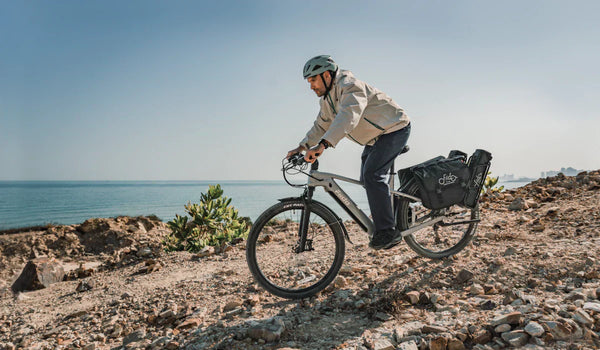 A rider takes a Fiido Nomads touring e-bike equipped with rear panniers across a rocky coastal trail.