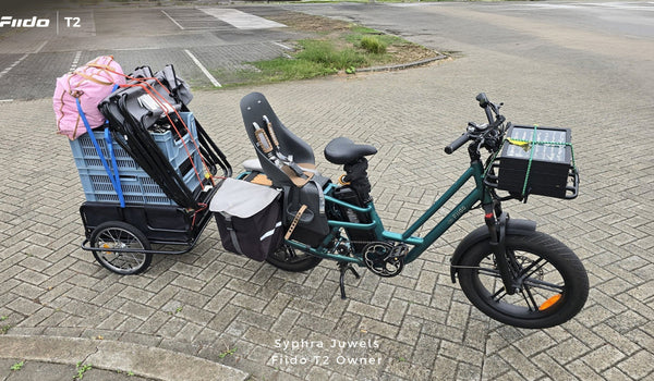 Un vélo cargo électrique Fiido T2 est présenté entièrement chargé, avec un siège enfant, des sacoches latérales, une caisse de transport avant et une remorque arrière lourdement chargée.