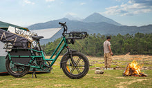 Vélo cargo électrique Fiido T2 chargé de matériel de camping sur un site de camping en montagne, à côté d’une tente et d’un feu de camp