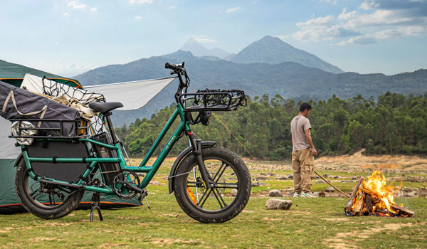 Vélo cargo électrique Fiido T2 chargé de matériel de camping sur un site de camping en montagne, à côté d’une tente et d’un feu de camp